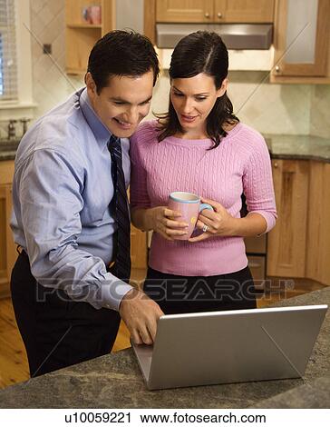 Stock Image - Mid-adult couple looking at laptop computer while standing in kitchen.. Fotosearch