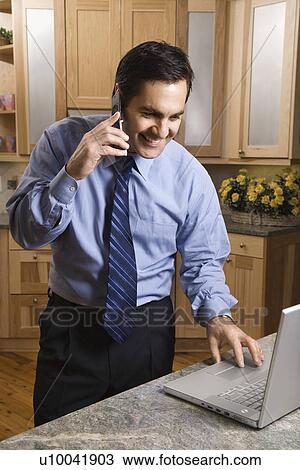 Stock Image - Mid-adult male talking on cellphone and looking at laptop computer in kitchen.. Fotosearch