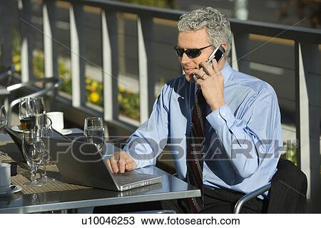 Stock Image - Prime adult Caucasian man in suit sitting at patio table outside looking at laptop and talking on cellphone.. Fotosearch