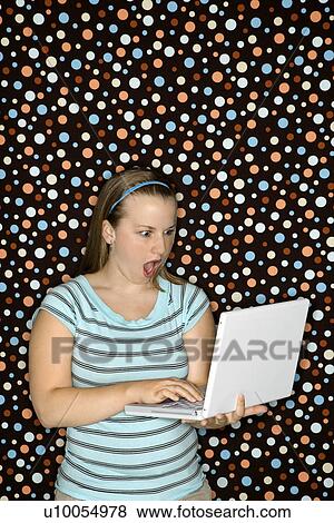 Stock Photo - Young Caucasian woman typing on laptop and looking shocked.. Fotosearch