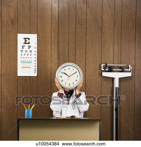 Caucasian male doctor sitting at desk holding clock over face. View Large Photo Image Picture - Caucasian male doctor sitting at desk holding clock over face.. Fotosearch