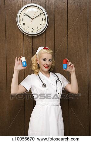 Stock Image - Caucasian mid-adult female nurse standing by wood paneling holding oversized pills and smiling at viewer.. Fotosearch