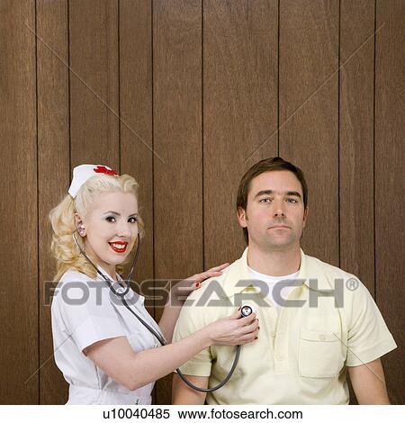Stock Photography - Caucasian mid-adult female nurse checking male's heartbeat by wood paneling.. Fotosearch
