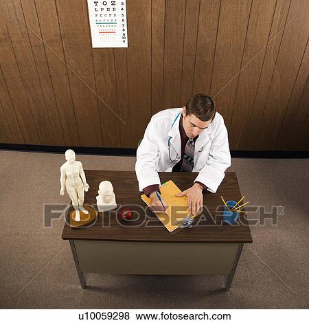 Stock Photo - High angle of mid-adult Caucasian male doctor at desk writing.. Fotosearch