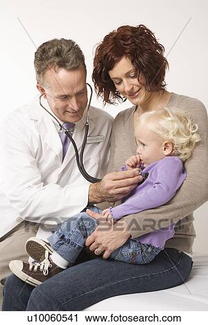 Stock Image - Middle-aged adult Caucasian male doctor holding stethoscope to female toddler's chest with mother watching.. Fotosearch