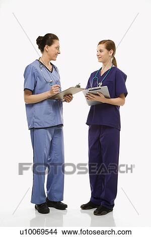 Picture - Portrait of Caucasian women doctors in medical scrubs standing talking holding medical charts against white background.. Fotosearch