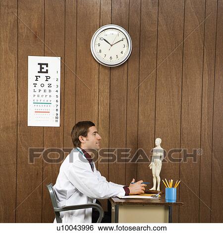 Stock Photograph - Side view of mid-adult Caucasian male doctor sitting at desk. . Fotosearch