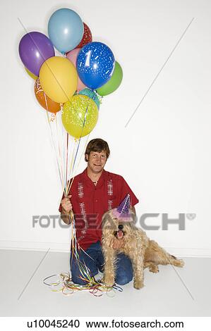 Stock Image - Caucasian young adult male sitting with brown fluffy dog wearing party hat with balloons.. Fotosearch