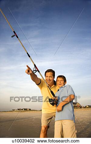 Stock Image - Caucasian mid-adult man shore fishing on beach with pre-teen boy and pointing.. Fotosearch