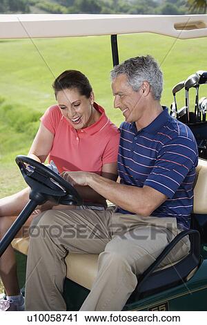 Stock Image - Mid-adult male and female in golf cart pointing at score card. . Fotosearch