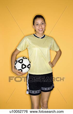 Stock Photograph - Portrait of smiling Multi-racial teen girl holding soccer ball at hip standing against yellow background.. Fotosearch