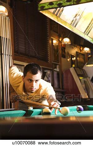 Stock Photo - Young man concentrating while aiming at pool ball while playing billiards.. Fotosearch