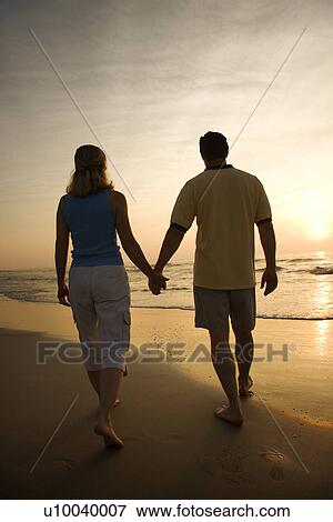 Stock Photo - Caucasian mid-adult couple walking holding hands on beach at sunset.. Fotosearch
