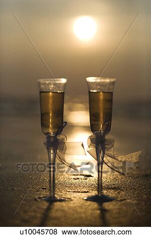 Stock Photo - Pair of flute glasses filled with champagne with bows on stems on beach at sunset.. Fotosearch