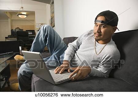 Stock Photograph - Asian young adult man wearing backwards baseball cap lounging on sofa working on laptop and looking at viewer.. Fotosearch