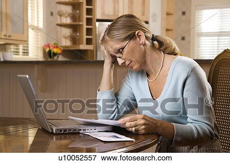 Stock Photo - Caucasian mid-adult woman paying bills on laptop computer resting head in hand with worry.. Fotosearch
