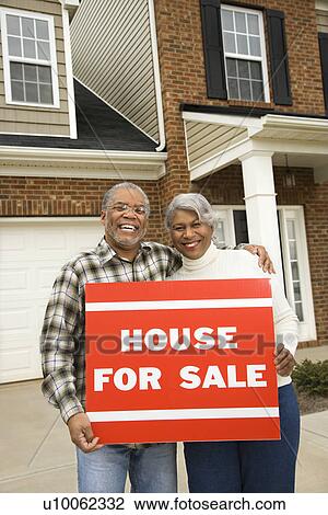 Stock Image - Portrait of middle-aged African-American couple outside house with for sale sign.. Fotosearch