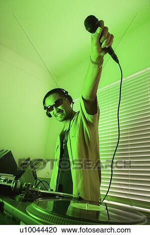 Stock Image - Smiling Asian young adult male DJ wearing sunglasses and holding microphone up in front of mixing equipment and turntable.. Fotosearch