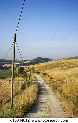 Stock Image - Telephone poles beside dirt road leading through rolling hills in countryside of Tuscany, Italy.. Fotosearch