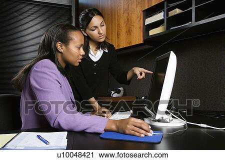 Stock Image - African-American and Indian young adult business women looking and pointing at computer in office.. Fotosearch