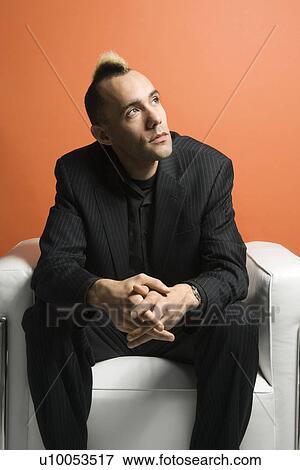 Stock Photo - Caucasian man with mohawk wearing suit sitting in chair against orange background.. Fotosearch