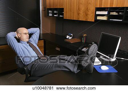 Stock Image - Caucasian middle-aged businessman in office sitting with feet on desk.. Fotosearch