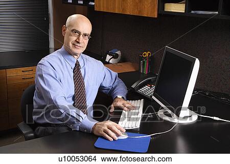 Caucasian middle-aged businessman sitting at desk in office in front of computer. View Large Photo Image Picture - Caucasian middle-aged businessman sitting at desk in office in front of computer.. Fotosearch