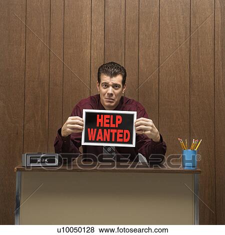 Stock Photo - Caucasion mid-adult retro businessman with desperate expression sitting at desk holding up help wanted sign.. Fotosearch