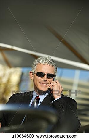 Stock Image - Close up of prime adult Caucasian man in suit sitting at patio table outside talking on cellphone and smiling.. Fotosearch