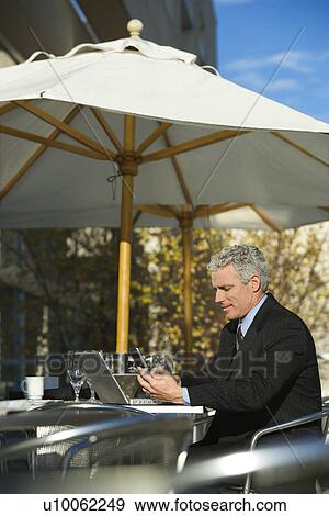 Stock Photo - Prime adult Caucasian man in suit sitting at patio table outside with laptop and dialing cellphone.. Fotosearch