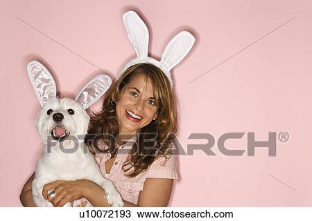 Stock Image - Caucasian prime adult female and white terrier dog wearing rabbit ears.. Fotosearch