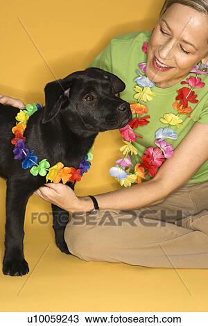 Stock Image - Caucasian prime adult female sitting with black puppy wearing leis.. Fotosearch