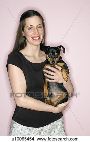 Smiling Caucasian woman holding Miniature Pinscher dog standing against pink background. View Large Photo Image Picture - Smiling Caucasian woman holding Miniature Pinscher dog standing against pink background.. Fotosearch
