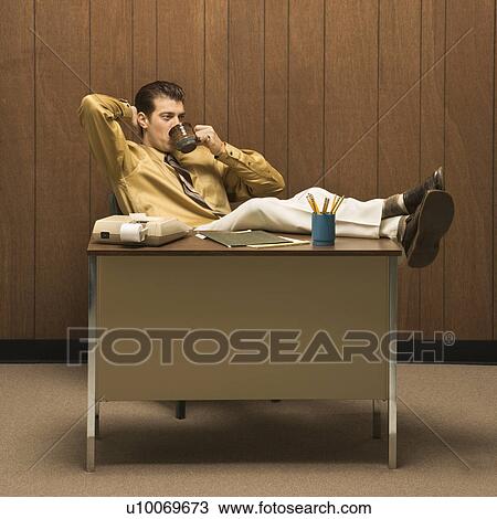 Stock Image - Caucasion mid-adult retro businessman sitting with feet propped on desk leaning back with hand behind head drinking coffee.. Fotosearch