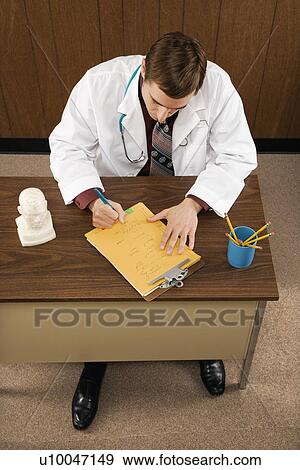Stock Photo - High angle of mid-adult Caucasian male doctor at desk writing.. Fotosearch