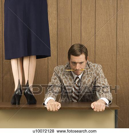 Stock Photo - Mid-adult Caucasian male biting pencil and holding desk with Caucasian female standing on desk beside him.. Fotosearch