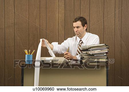 Stock Image - Mid-adult Caucasian male holding printout from calculator in office setting.. Fotosearch