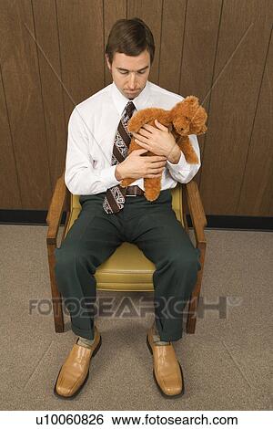 Stock Photograph - Mid-adult Caucasian male sitting in chair holding a stuffed animal looking sad.. Fotosearch