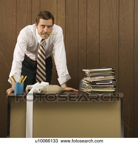 Stock Image - Mid-adult Caucasian male with pencil in his mouth and hands on a desk.. Fotosearch