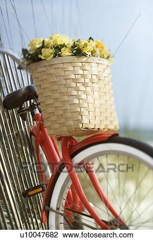 Stock Image - Red vintage bicycle with basket and flowers leaning against wooden fence at beach.. Fotosearch