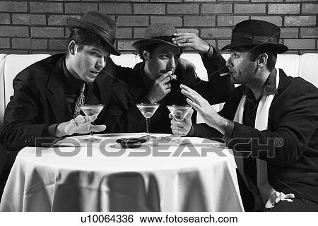 Stock Photograph - Three Caucasian prime adult males in retro suits sitting at table drinking and smoking and talking.. Fotosearch