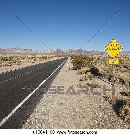 Stock Image - Road in desert with sign for soft shoulder and mountains in distance.. Fotosearch