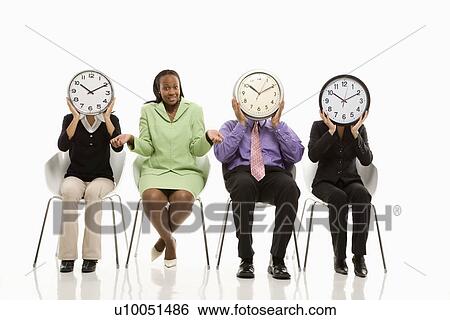 Stock Photograph - Business people sitting holding clocks over faces while African-American businesswoman shrugs.. Fotosearch