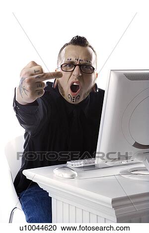 Stock Image - Caucasian mid-adult man with tattoos and piercings using computer holding up middle finger.. Fotosearch