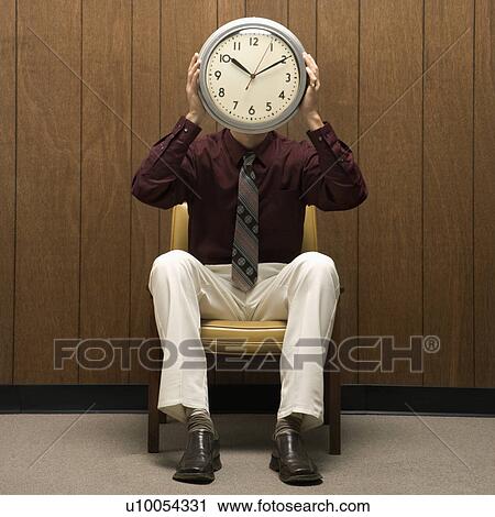 Stock Image - Caucasion mid-adult retro businessman sitting in chair against wood paneling holding clock over face.. Fotosearch