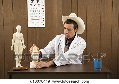 Stock Photo - Mid-adult Caucasian male doctor wearing cowboy hat sitting at desk playing with figurine.. Fotosearch
