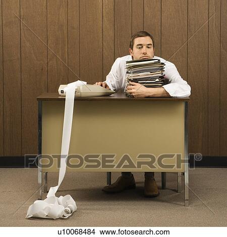 Picture - Mid-adult Caucasian male holding stack of paperwork with hand on calculator with long printout.. Fotosearch
