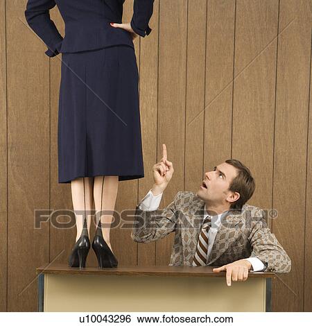 Stock Photograph - Mid-adult Caucasian male pointing up to Caucasian female standing on desk.. Fotosearch