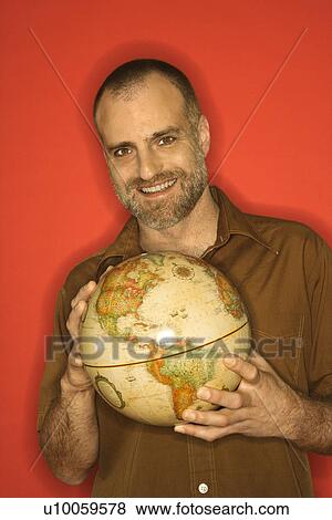Stock Photo - Portrait of smiling Caucasian man holding globe standing against orange background.. Fotosearch