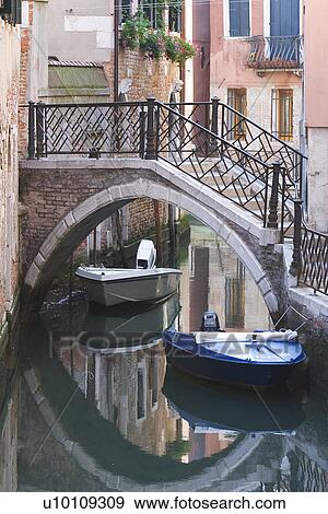 Stock Photo - Building reflected in quite canal - boats moored at side. Bridge with steps over canal.. Fotosearch